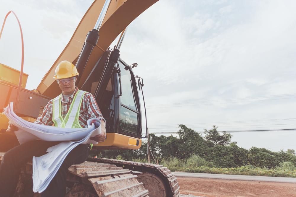 Worker in a hard hat reviewing plans beside an excavator