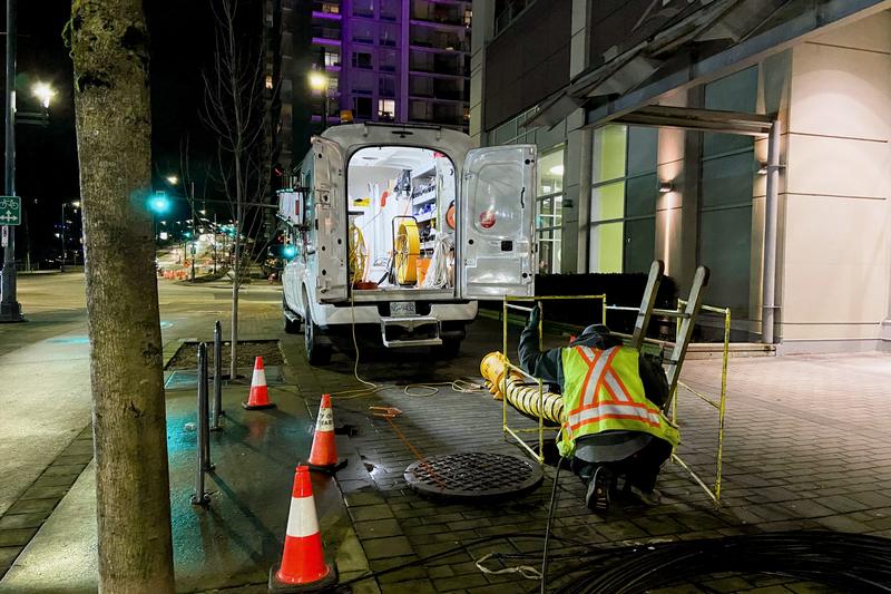 Utility van parked at night beside an open manhole with a guard rail, ventilation hose and traffic cones